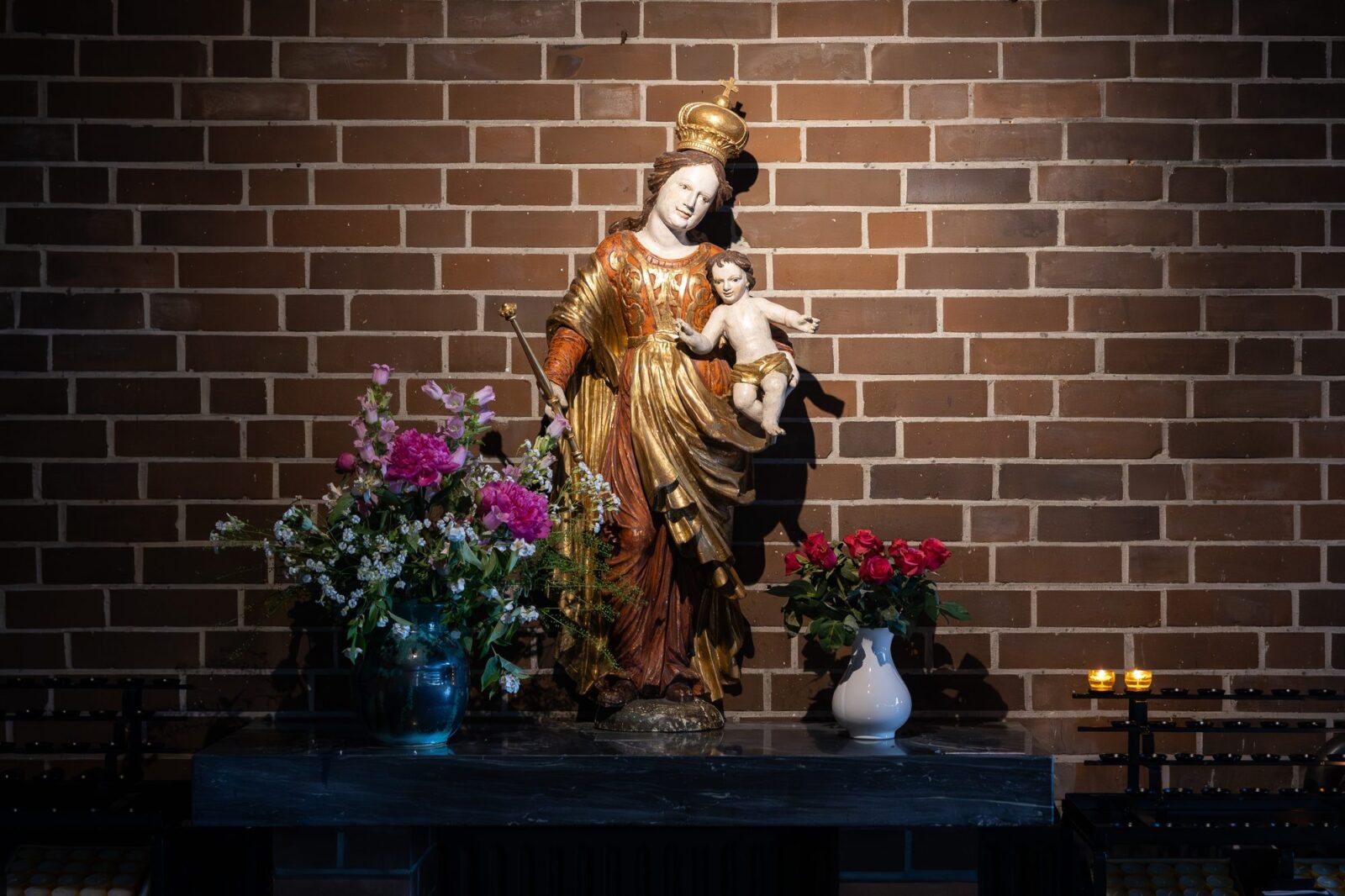 Madonna mit Kind vor der Backsteinwand, davor Blumen und Kerzen